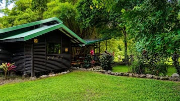 A wooden cabin with a green roof, surrounded by lush greenery and a well-maintained lawn.