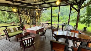 An outdoor patio with wooden furniture, including a sofa, chairs, and a table. There is a hot tub and a grill in the background. - Cabaña Los Tucanes - Paradise for Couples! (La Fortuna, Costa Rica)