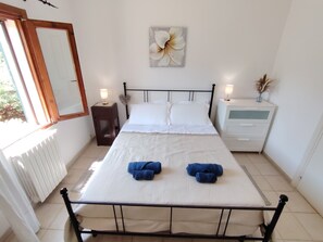 A neatly made bed with white linens and two blue towels, a bedside table with a lamp, a nightstand with a vase, and a wall-mounted flower painting.
