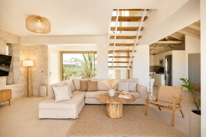 A modern living room with a sectional sofa, a round coffee table, a wicker chair, a staircase, and a kitchen area.