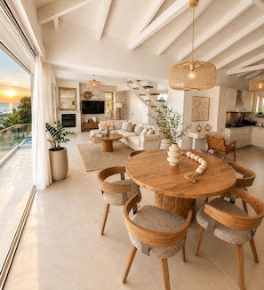 A spacious living area with a round wooden table, cushioned chairs, a sofa, and a staircase leading to an upper level.