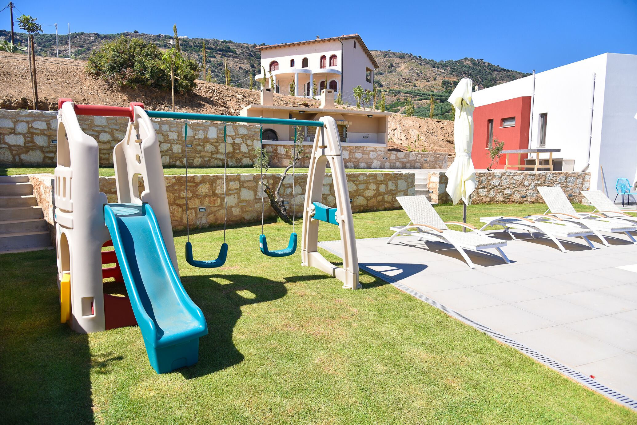 A playground with slides and swings, a pool area with lounge chairs, and a building with a red and white facade.