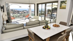 Ein modernes Wohnzimmer mit großem Fenster, einer Couch, einem Esstisch mit Früchten und Blick aufs Meer.