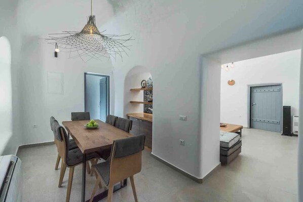 A modern dining area with a wooden table and chairs, a unique hanging light fixture, and a view into another room with a bed and a wardrobe.