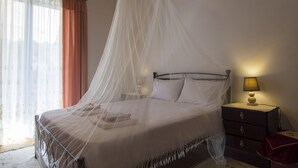 A bedroom with a bed covered in a white canopy, a nightstand with a lamp, and a window with curtains. - Cosi Apartment in village (Lakka Paxos)