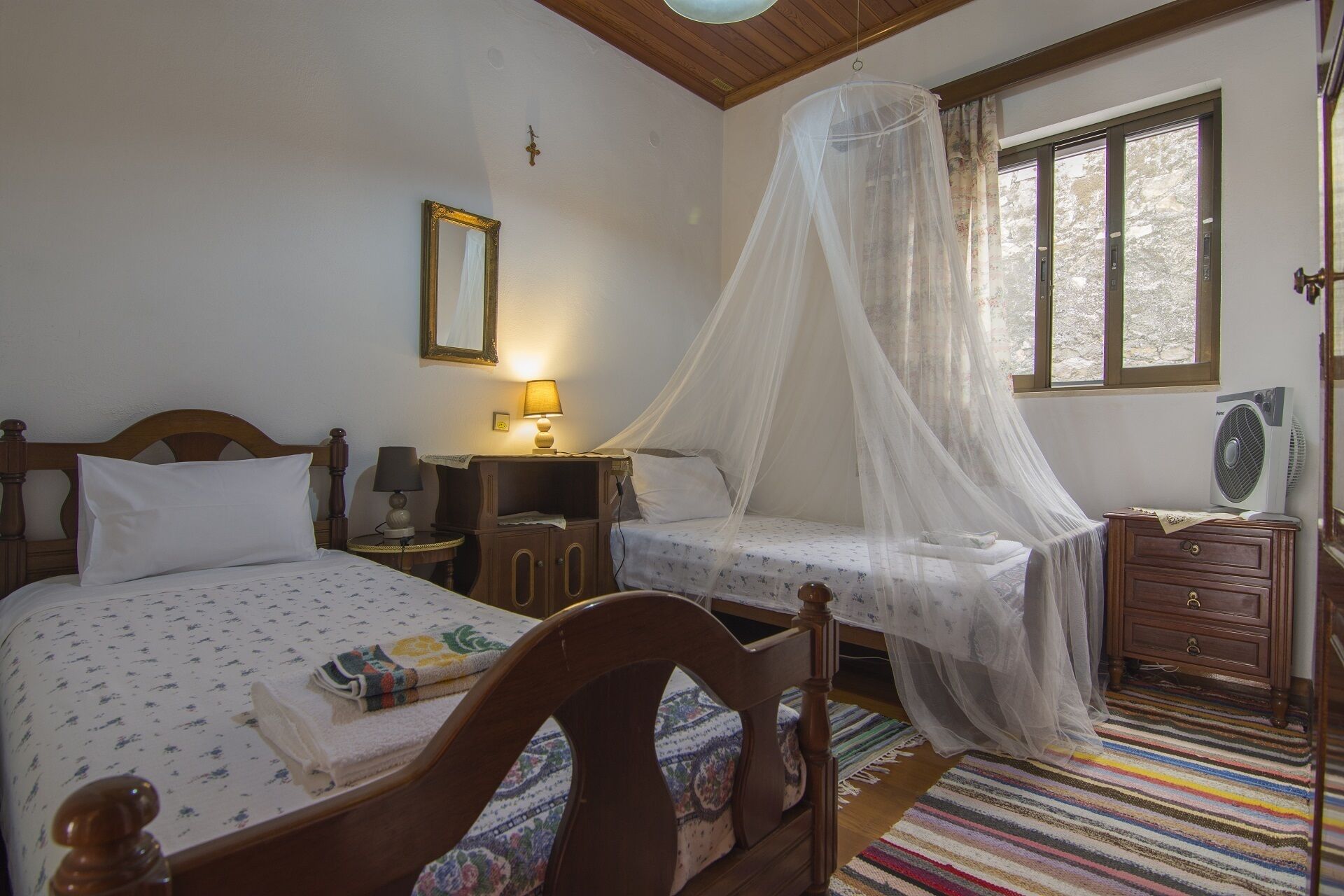 A bedroom with a bed, bedside table, lamp, bedside mirror, window, and a fan.