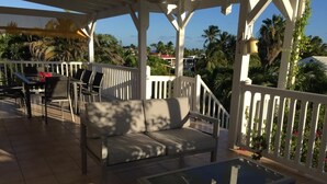 Een overdekt terras met een bank en een glazen tafel, omringd door een witte balustrade en palmbomen op de achtergrond.