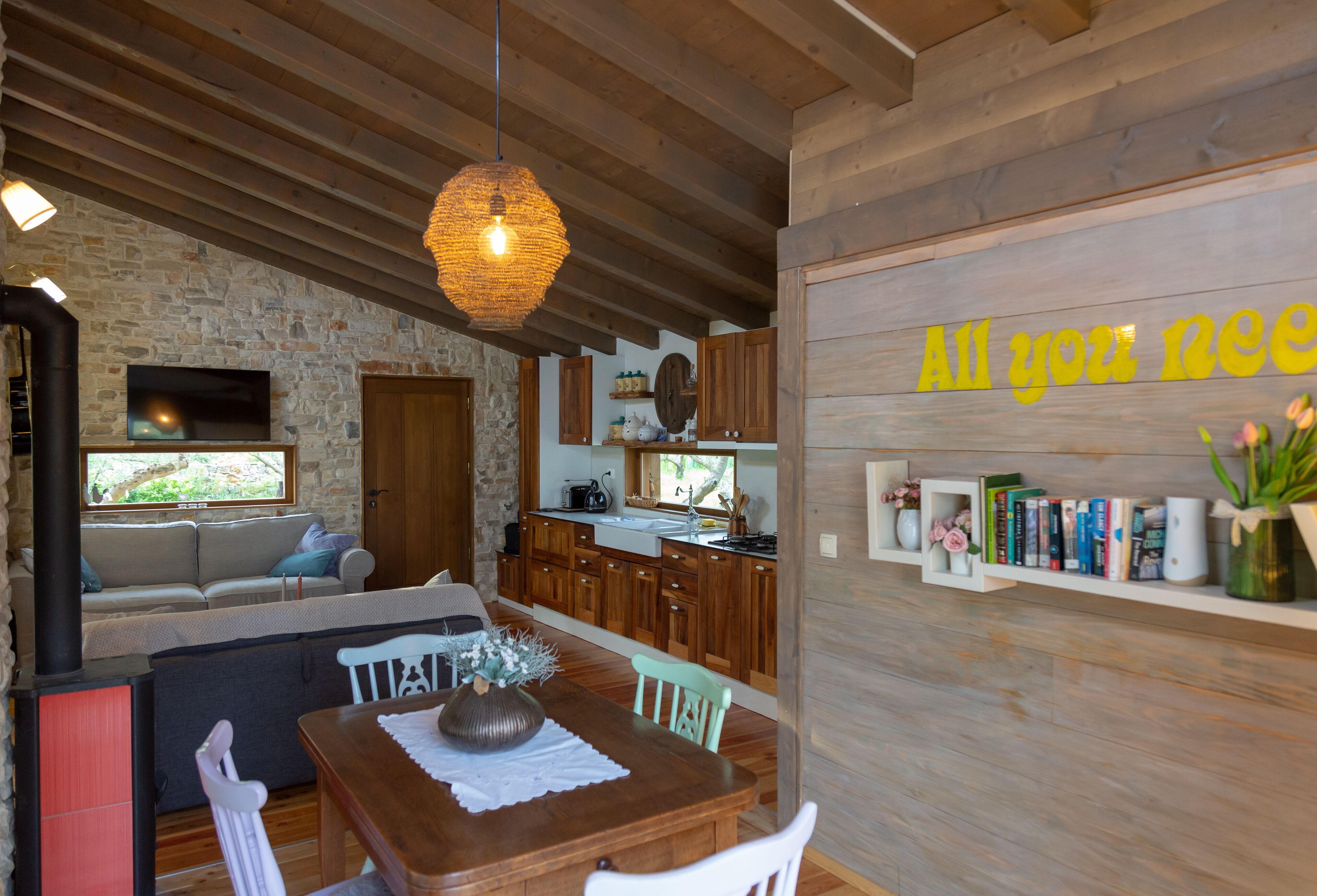 A cozy interior with a stone wall, wooden ceiling, a dining area, kitchen, and a shelf with books and flowers.