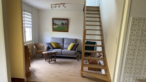A living room with a wooden staircase, a grey sofa with cushions, a wicker chair, and a wooden table with a laptop. - Quiet location, ideal for relaxing, house 5 km from the beaches of Binic-Etables (Lantic)