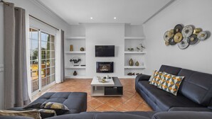 Un salon moderne avec un canapé, une table basse et un téléviseur. On y trouve des étagères ornées d’objets décoratifs et une grande fenêtre avec des rideaux.
