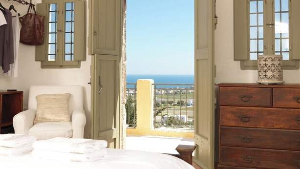 Une chambre avec un lit, une chaise, une commode et une vue sur la mer, visible à travers des volets ouverts.