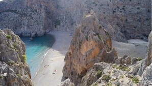 A rocky coastal landscape with a narrow beach and clear blue waters.