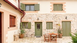 A stone building with a courtyard, featuring a door, windows with shutters, a table with chairs, and potted plants.