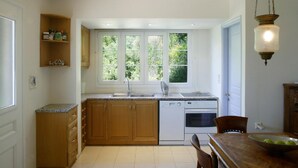 A kitchen with wooden cabinets, a white dishwasher, a sink, and a window with a view of greenery. - Villa Mimosa - Agios Nikitas Villas (Άγιος Νικήτας)