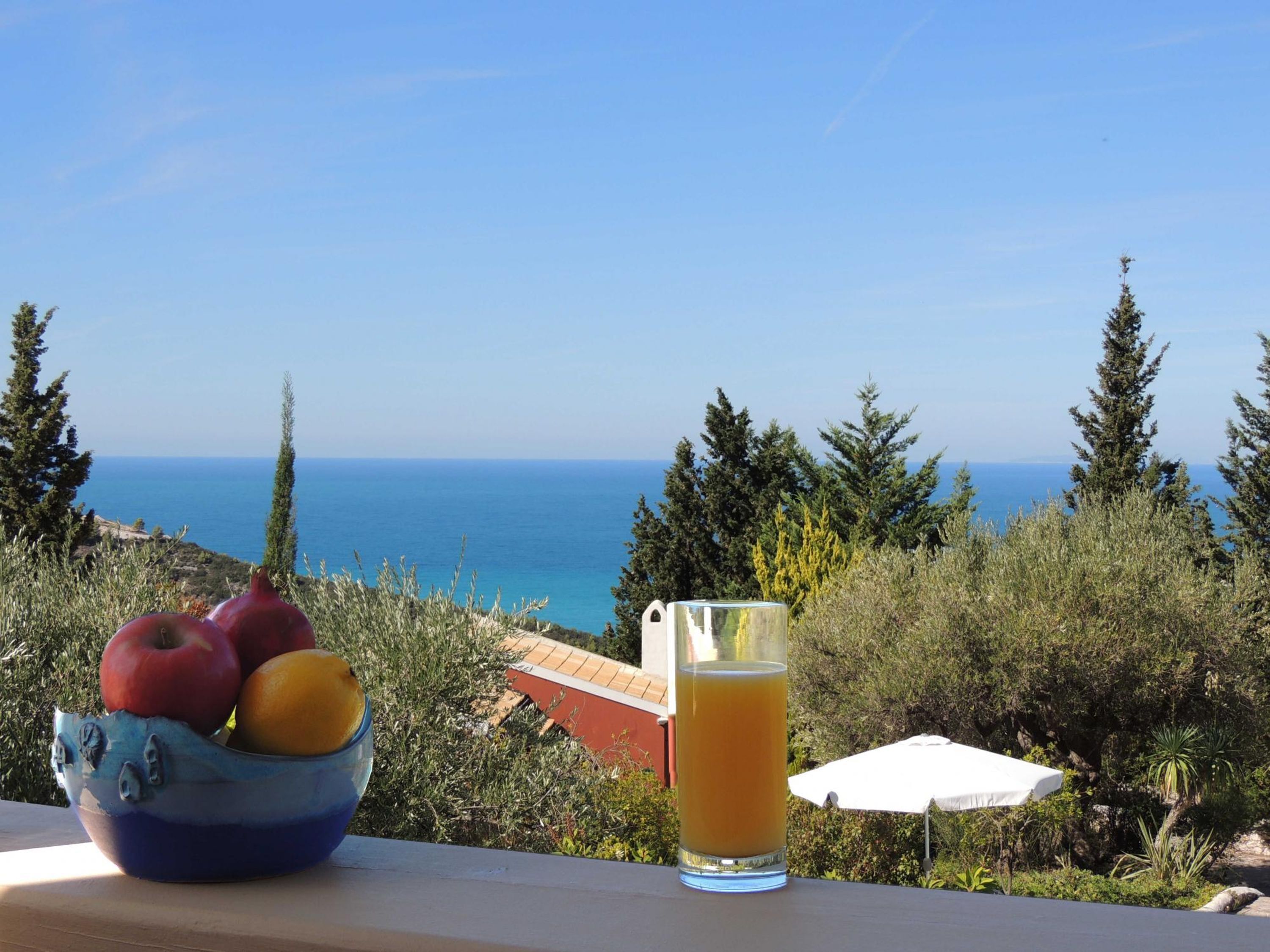 Mesa com uma fruteira contendo maçãs e laranjas, e um copo de suco de laranja, com vista para a costa.