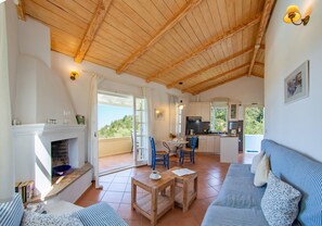 A spacious living area with a fireplace, a dining table, and a sofa. There is a kitchen in the background with a view of greenery. - Apartment Ilex - Agios Nikitas Villas (Άγιος Νικήτας)