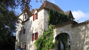 A stone building with red shutters, a car parked in front, and a large archway. - Enchanting Château with en-suites, Pool, Tennis, Grand Piano, BBQ, Fast WiFi (Thézac)
