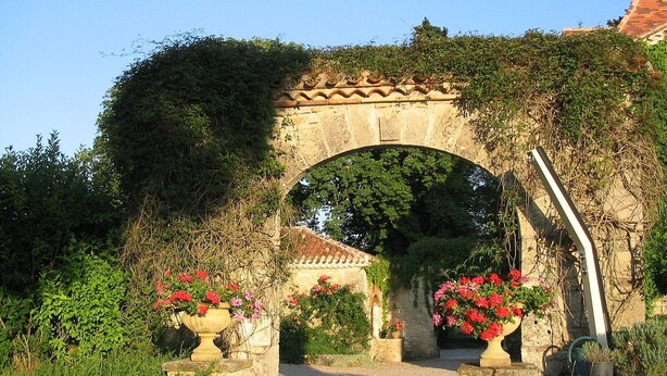 An arched stone gateway with flowering plants on either side, leading to a building with a tiled roof. - Enchanting Château with en-suites, Pool, Tennis, Grand Piano, BBQ, Fast WiFi (Thézac)