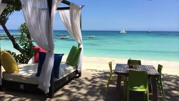 Relax under a shaded canopy, with a comfortable bed and dining area overlooking the beach and turquoise waters.