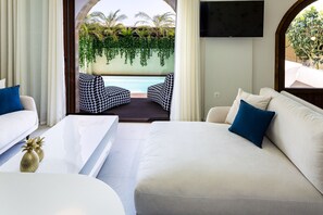 A modern living room with a white sofa, a coffee table, and patterned chairs. A TV is mounted on the wall, and there is a view of a pool and greenery outside.