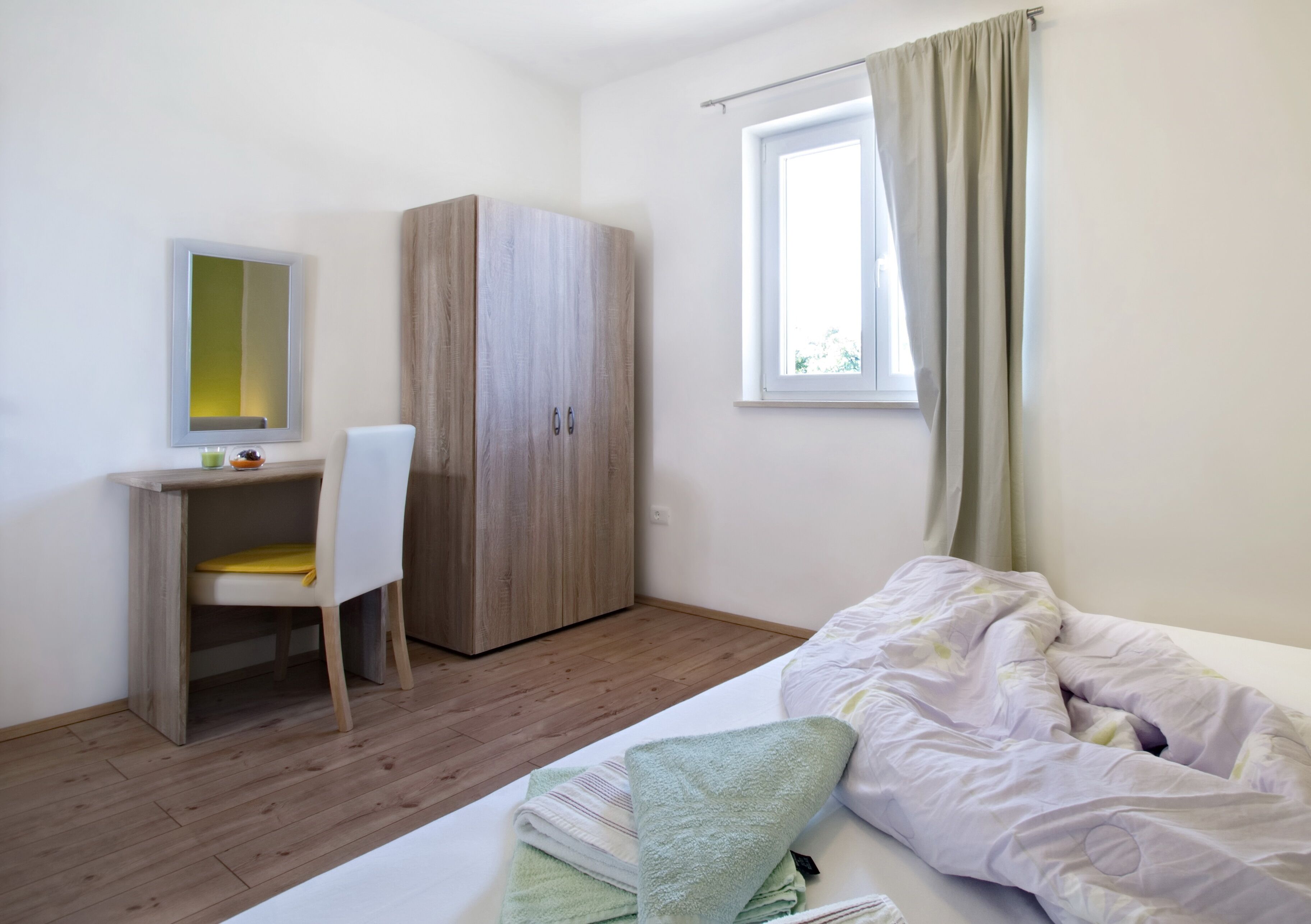 A bedroom with a bed, a desk with a chair, a wardrobe, a window with curtains, and a mirror on the wall.