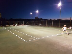 Ein Tennisplatz im Freien bei Nacht, beleuchtet mit hellem Licht, inklusive Netz und Umzäunung.
