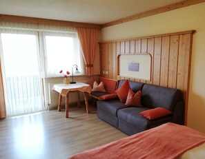 A cozy room with a wooden floor, a grey sofa with red cushions, a small table with a lamp, and a window with sheer curtains.
