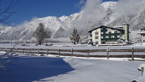 Un paysage montagneux enneigé, avec un bâtiment à la façade verte et blanche, probablement un hôtel ou un chalet, sous un ciel dégagé et des arbres couverts de neige.