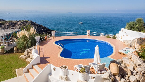 A pool with a curved edge, surrounded by white walls and a stone deck. There are lounge chairs and a small umbrella. The pool overlooks a rocky coastline with a clear blue sea and a boat in the distance. - Villa Phaedra 7 bedroom villa (Tersanas)