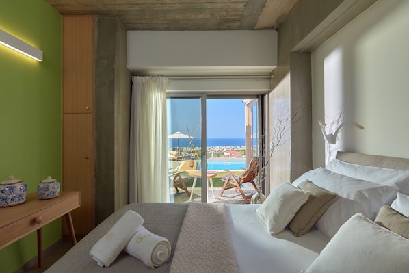 A hotel room with a bed, a wooden nightstand, a view of a pool and the sea, and a balcony with a chair and umbrella.