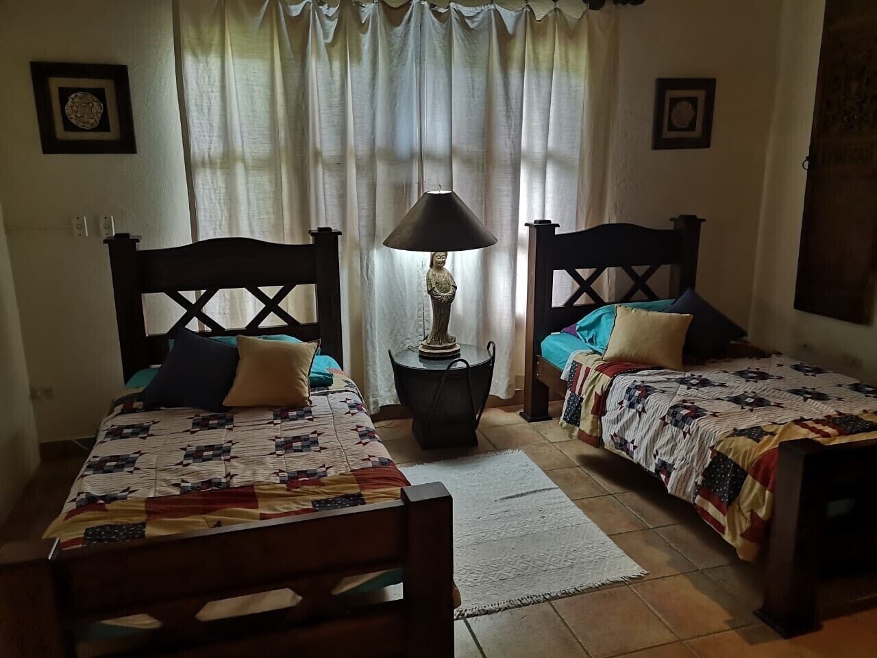 Une pièce avec deux lits en bois, une table ronde avec une lampe et une fenêtre avec des rideaux.
