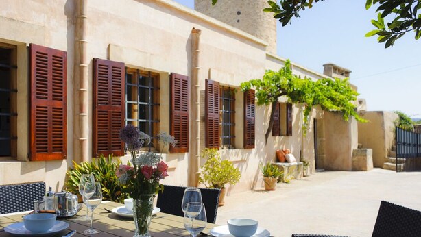 Un área de comedor al aire libre con mesas y sillas de madera, una pared de piedra y plantas en macetas.