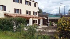 Un bâtiment à deux étages, doté d’un toit en tuiles, d’un patio aménagé avec du mobilier d’extérieur et d’un jardin où poussent différentes plantes, ainsi qu’un grand rocher.