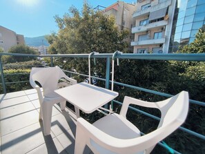 En balkon med hvide plaststole og et bord, med udsigt over en have og bygninger.