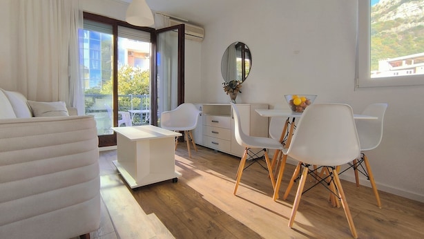 A modern, well-lit room with a sofa, a dining table with chairs, a small white cabinet, and a large window offering a view of the outdoors. - Bright 1BR apartment with two balconies, mountain views, sea glimpses and PKG (Будва)