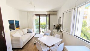 A modern living room with a sofa, dining table, chairs, and a view of the outdoors. - Bright 1BR apartment with two balconies, mountain views, sea glimpses and PKG (Будва)