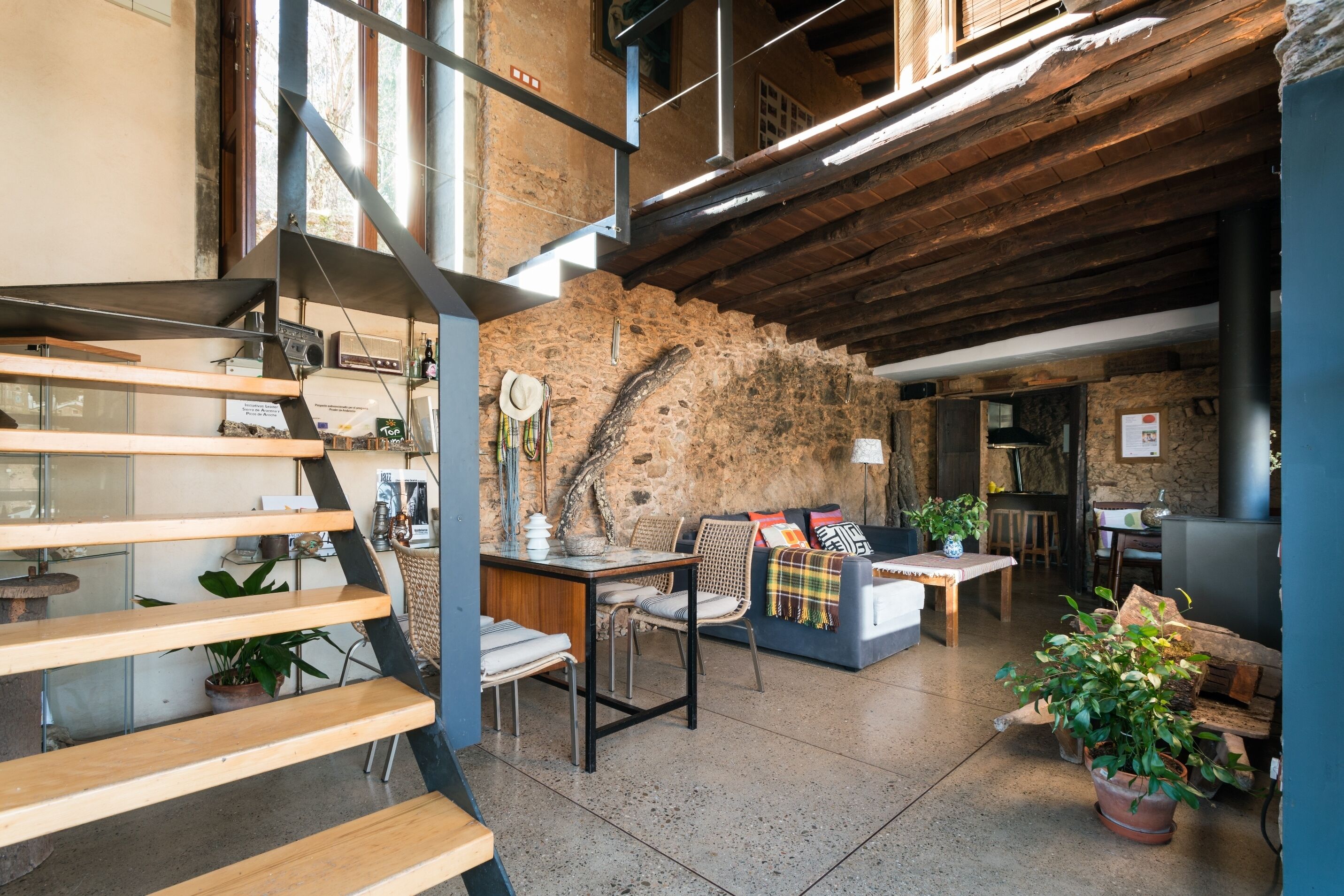 A rustic interior with exposed brick walls, a wooden staircase, a sofa with colorful cushions, and a dining area with a table and chairs.