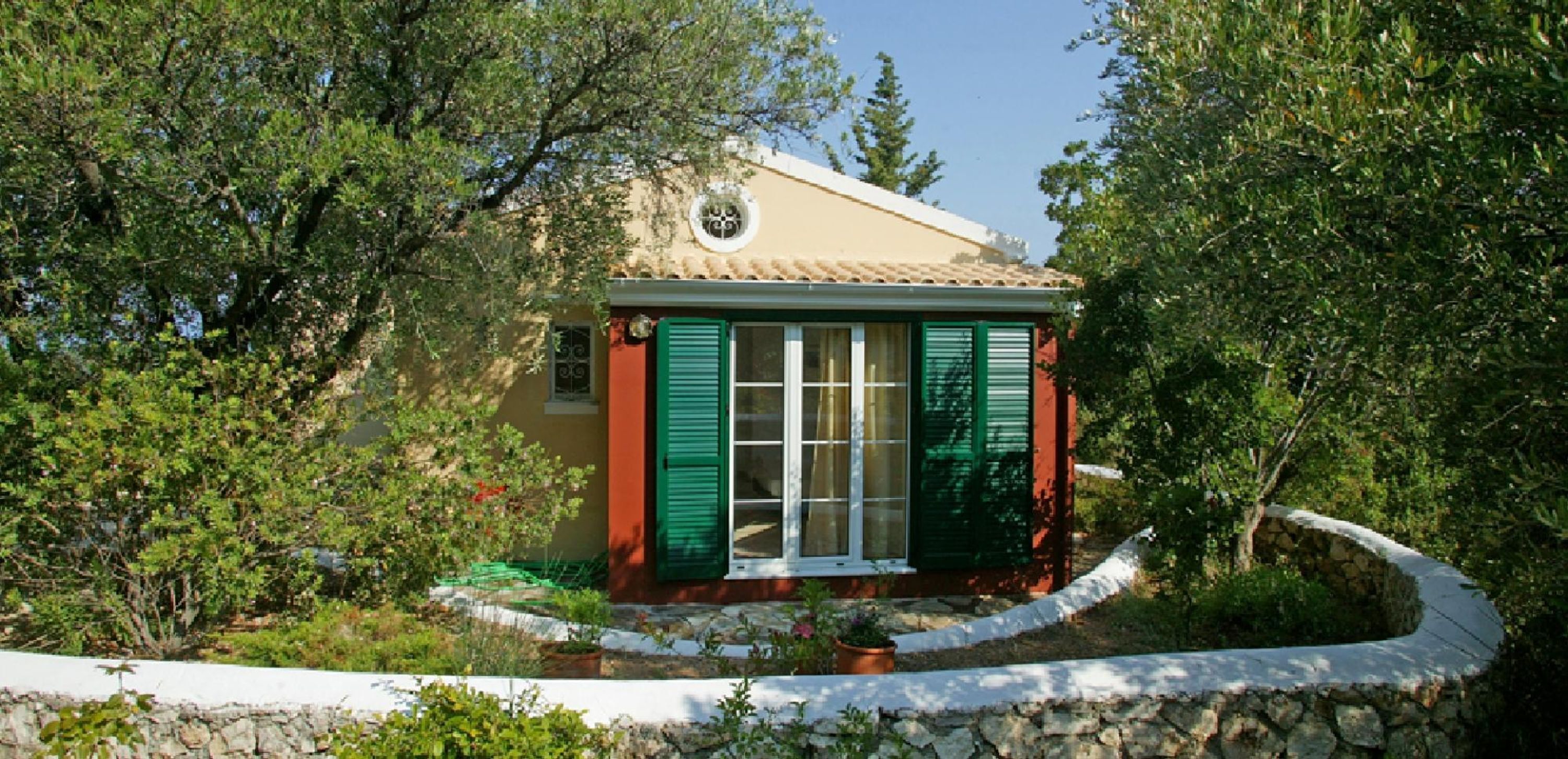 Una pequeña casa con un muro de piedra que rodea un jardín, con una ventana y contraventanas verdes y una puerta.