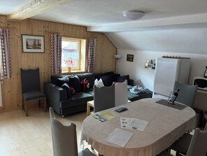 A cozy room with a wooden ceiling, a sofa, a dining table with papers, a refrigerator, and a window with curtains.