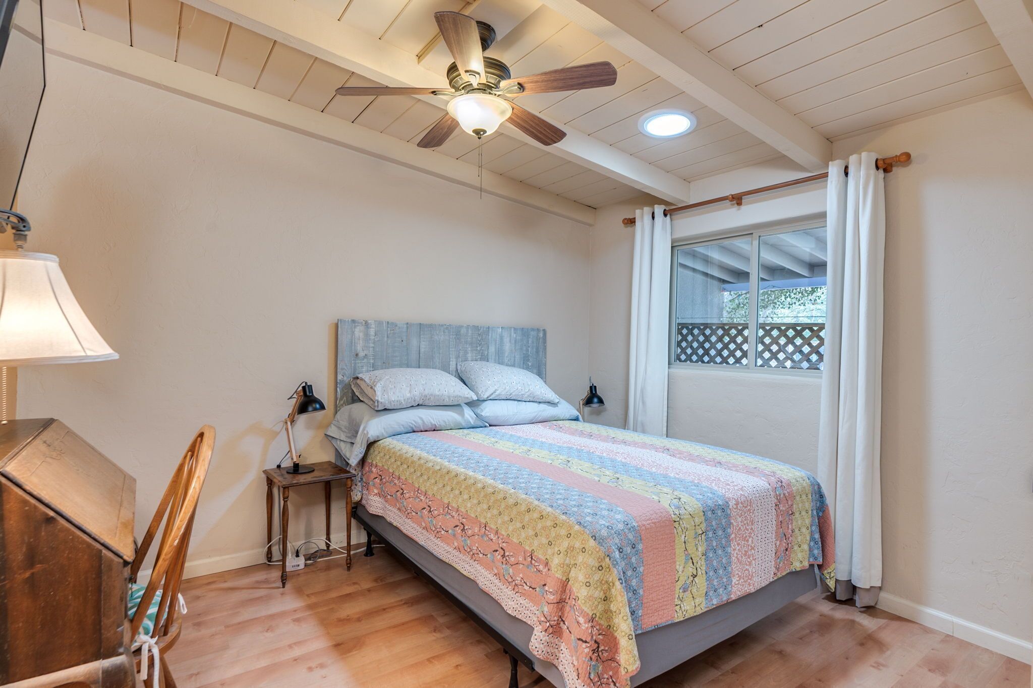 Un dormitorio con cama, ventilador de techo, ventana con cortinas, escritorio de madera y lámpara.