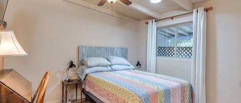 Un dormitorio con cama, ventilador de techo, ventana con cortinas, escritorio de madera y lámpara.