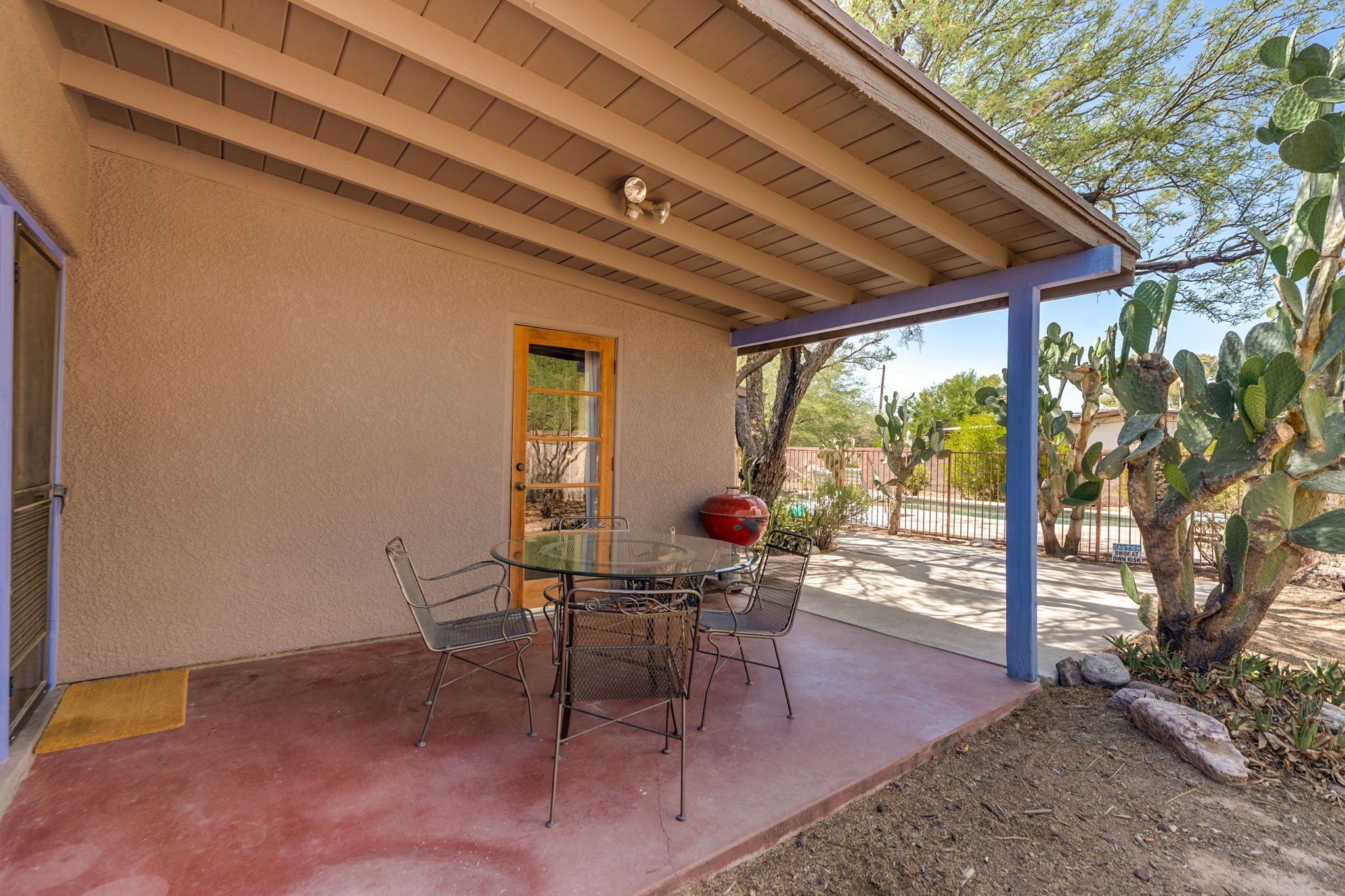 A covered patio with a round glass table and metal chairs, a wooden door, and a cactus plant.