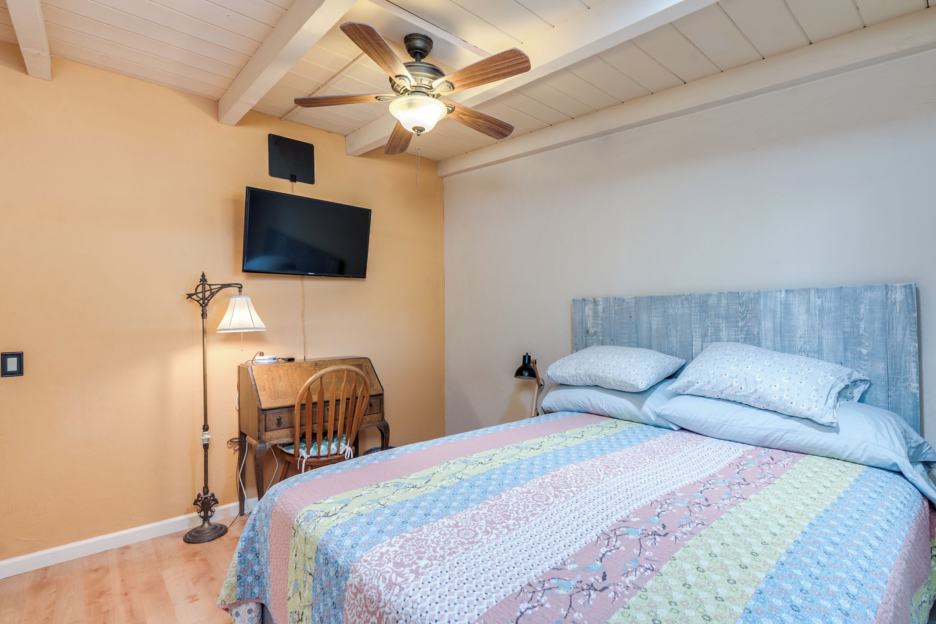 Un dormitorio con cama, ventilador de techo, escritorio, silla, lámpara y televisión.