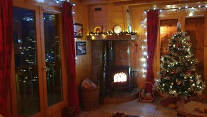 Une pièce chaleureuse avec une cheminée, un sapin de Noël et une fenêtre donnant sur un paysage urbain.