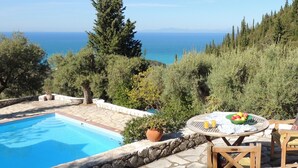 A poolside area with a view of the sea, surrounded by trees and a stone wall. - Villa Kiparissi - Agios Nikitas Villas (Άγιος Νικήτας)