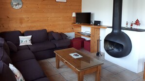 En hyggelig stue med trælofter, en sofa, et sofabord med glasplade, en pejs og et fjernsyn på en træhylde.