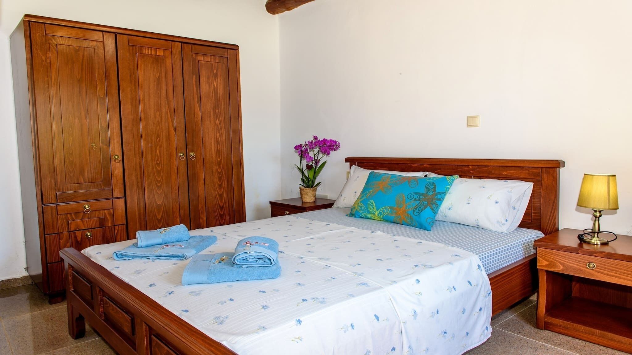 A bedroom with a wooden wardrobe, a bed with floral bedding, a nightstand with a lamp, and a potted orchid.