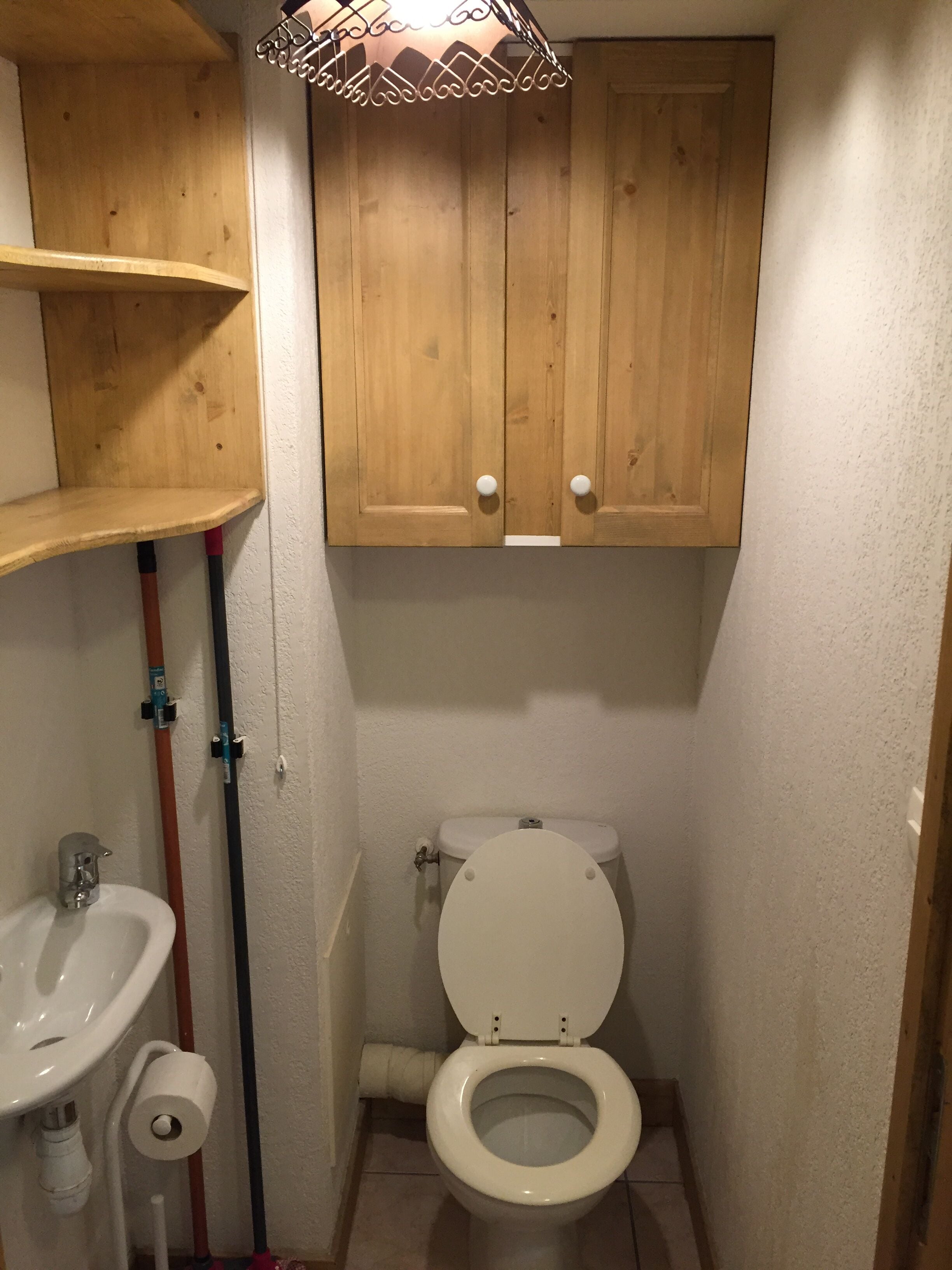 A small, compact bathroom with a wooden cabinet above the toilet, a sink with a faucet, and visible plumbing pipes.
