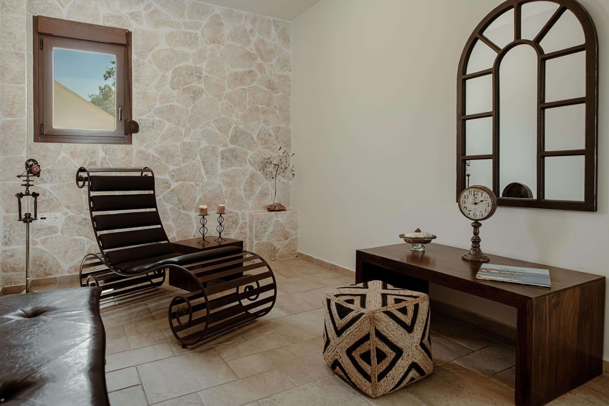 A room with a stone wall, a wooden-framed window, a black cushioned chair, a wooden side table with a clock, and a woven ottoman.