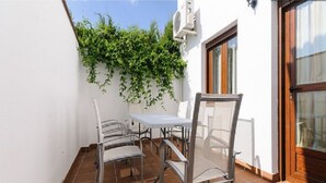 Une terrasse extérieure avec une table et des chaises, un sol carrelé, un mur blanc, une porte en bois et une fenêtre avec des rideaux.